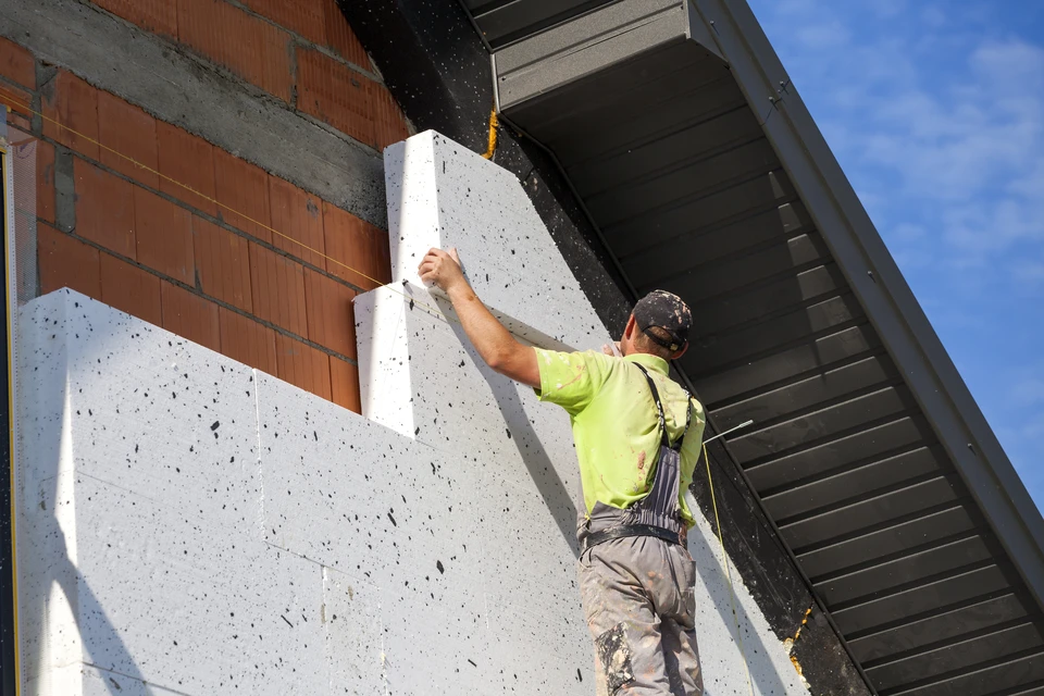 Chantier de construction extérieur avec ouvrier en combinaison bleue appliquant un enduit blanc sur mur béton